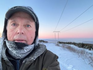 Charlie Sink wearing winter gear stands in a snowy landscape with frost on his face and clothing. The sky is a soft gradient of pink and blue, indicating sunrise or sunset. Power lines stretch into the distance, and a forested area can be seen in the background.