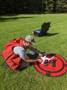 Clay Hoyt kneels on a grassy field, setting up a white drone on a red and black landing pad marked with the letter 'H'. He wears a red safety vest and a baseball cap. Behind him is an open black case containing equipment, and trees line the background on a sunny day.